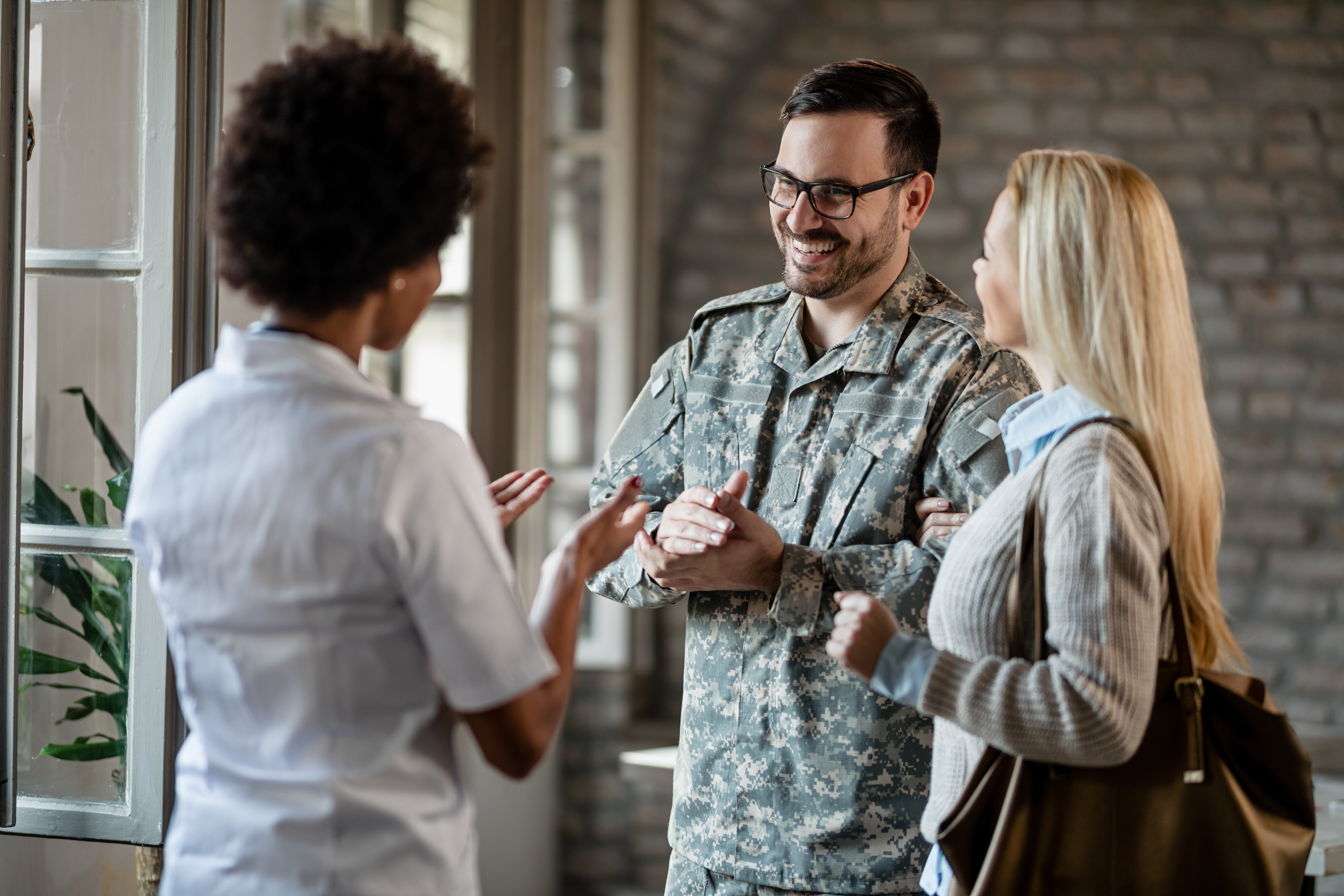 happy-army-soldier-his-wife-communicating-with-african-american-healthcare-worker-while-having-consultations-clinic